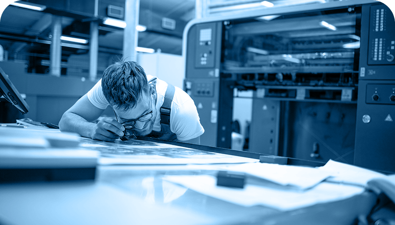 A worker examines a printed sheet beside industrial printing equipment