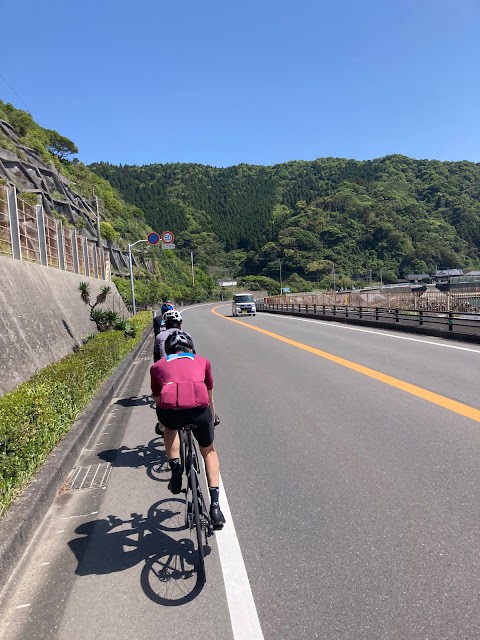Fellow cyclists riding along a sunny road beside wooded hills in Kyushu