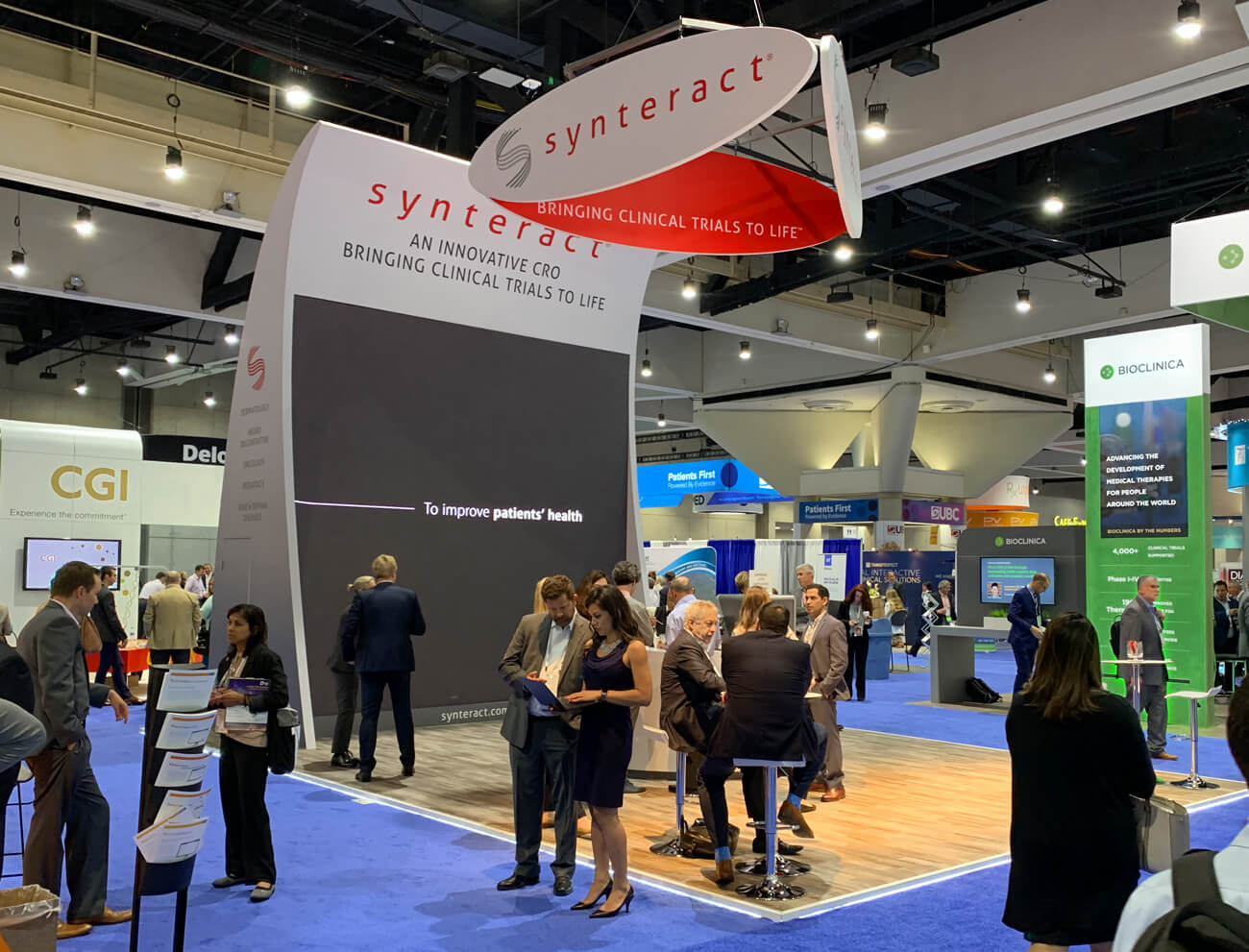 Visitors talking at the Synteract booth beneath suspended signage and a large screen