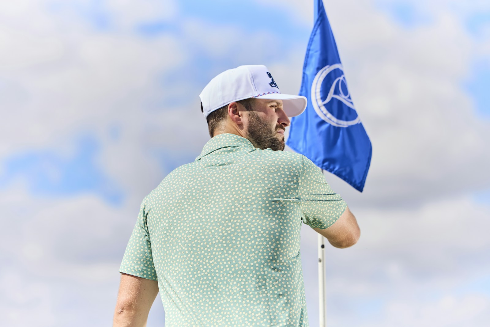 A golfer in a green patterned Breezy polo and white cap beside a blue golf flag