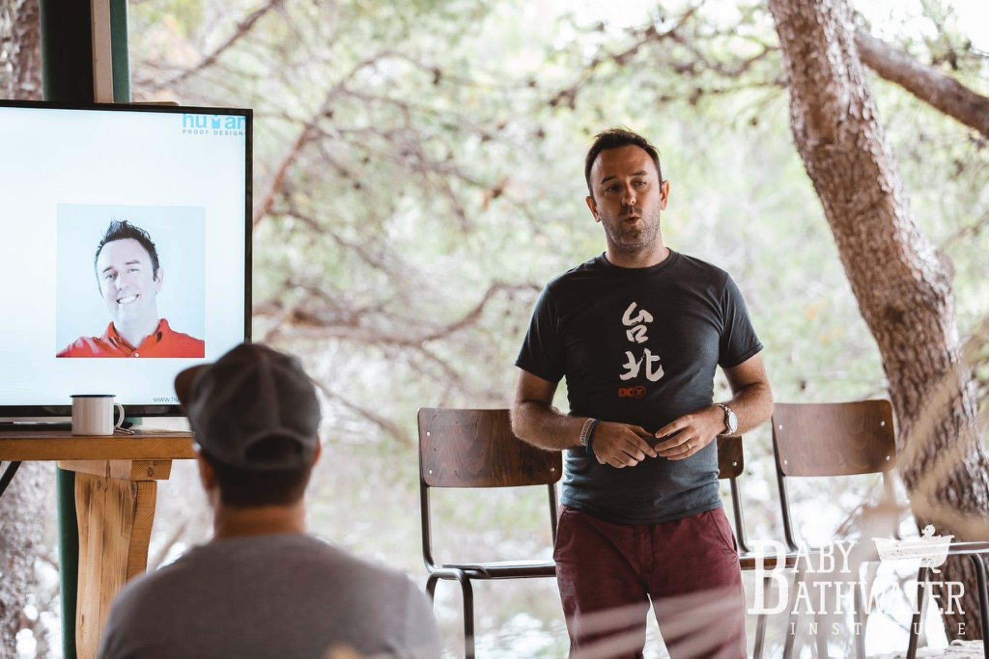 Dominic Wells speaking to an audience beside a Human Proof Designs presentation