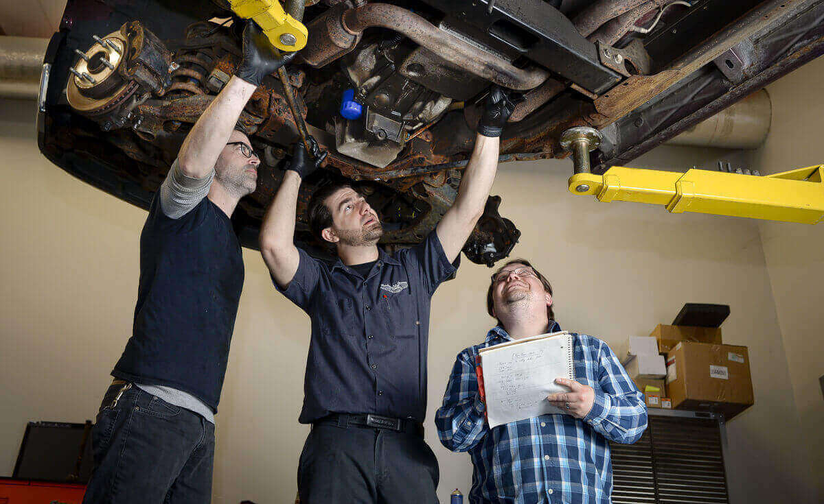 Three people studying the underside of a vehicle on a workshop lift during Dorman field research