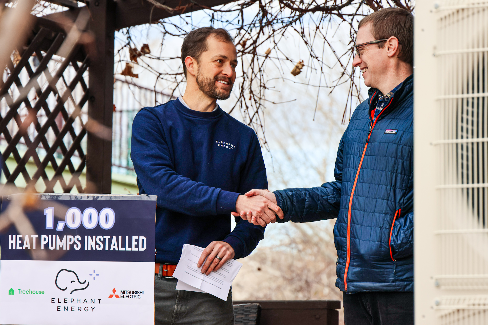 DR Richardson, left, shakes hands with homeowner Justin Bailey beside a sign celebrating 1,000 heat pumps installed.