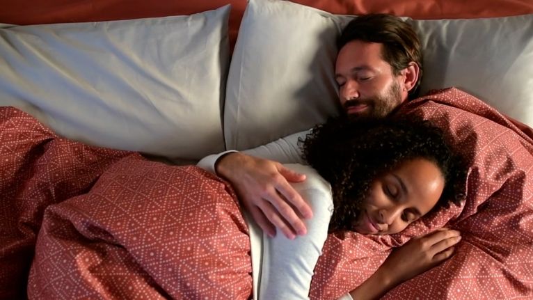 A couple tucked into red patterned bedding in a frame from DreamItReel's QuickZip advertisement