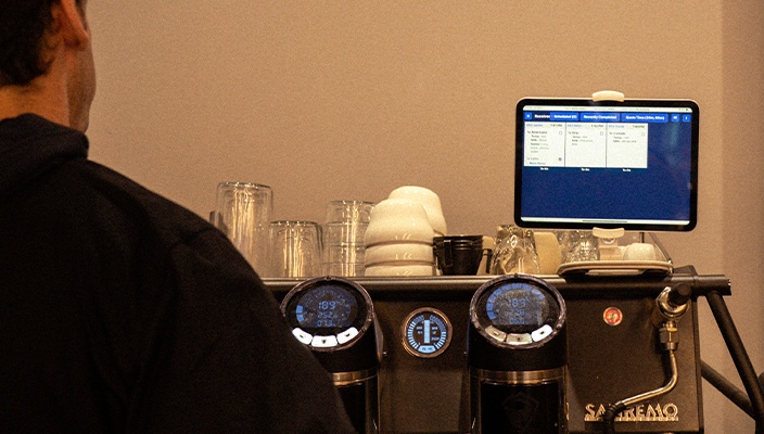 A barista beside an espresso machine with a Dripos ticket screen mounted above it