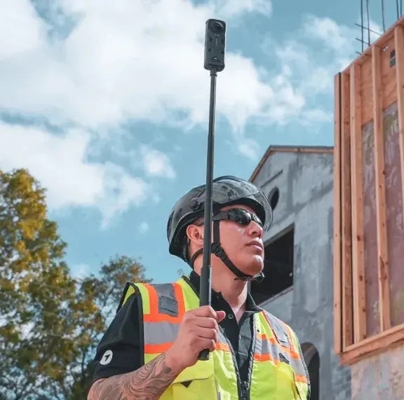 A construction worker carrying a pole-mounted 360-degree camera on a building site