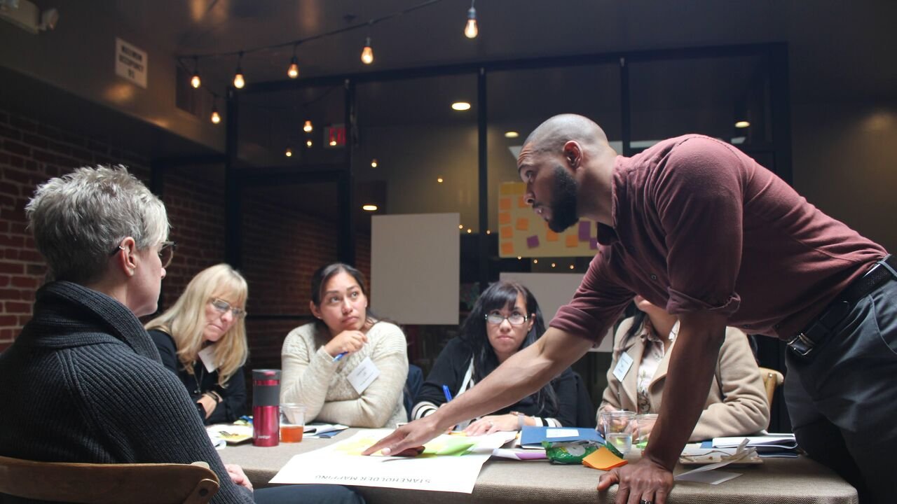 Durell Coleman leans across a workshop table with participants, papers and sticky notes.