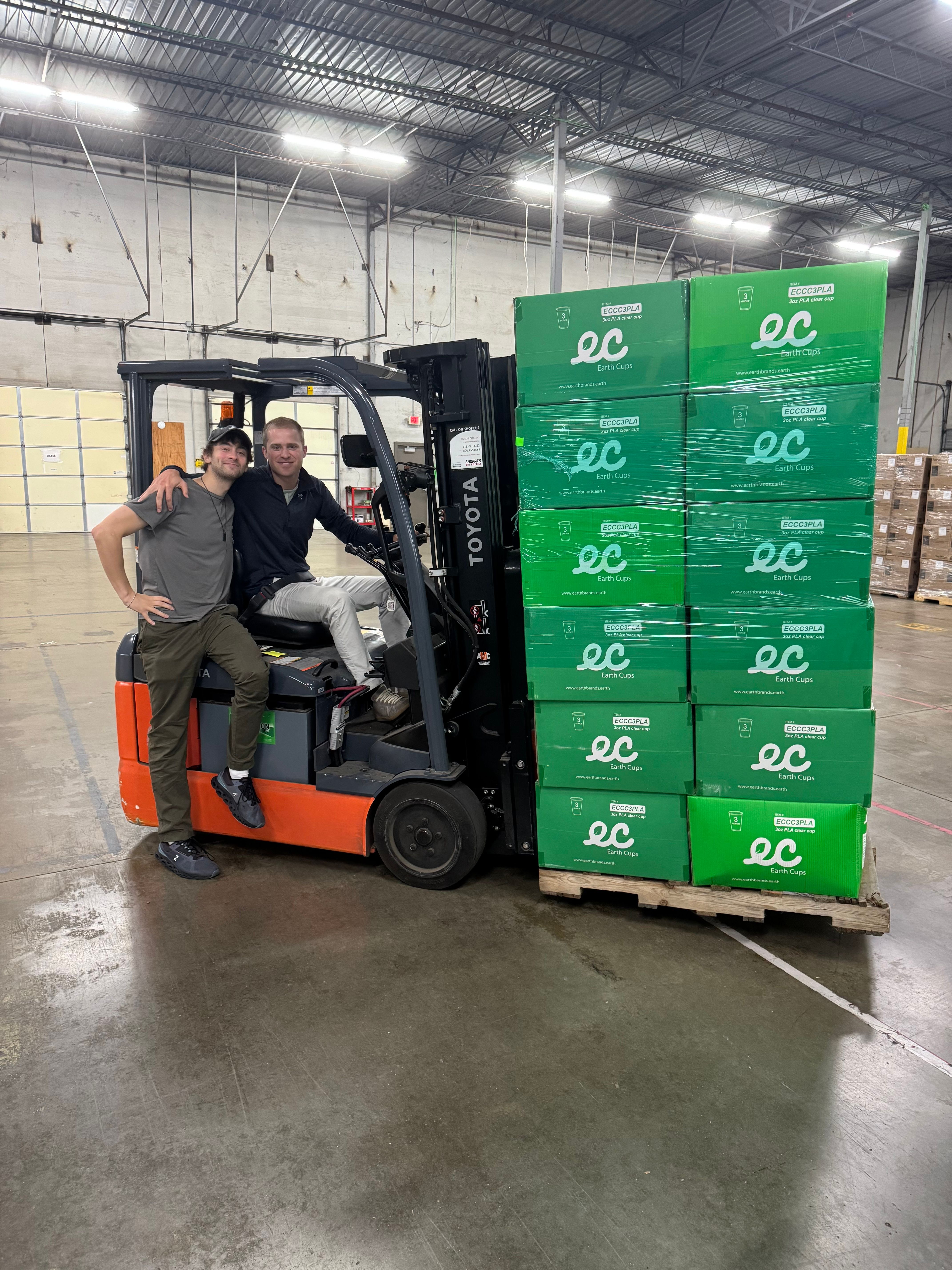 Earth Brands founders beside a forklift and a pallet of green Earth Cups cartons