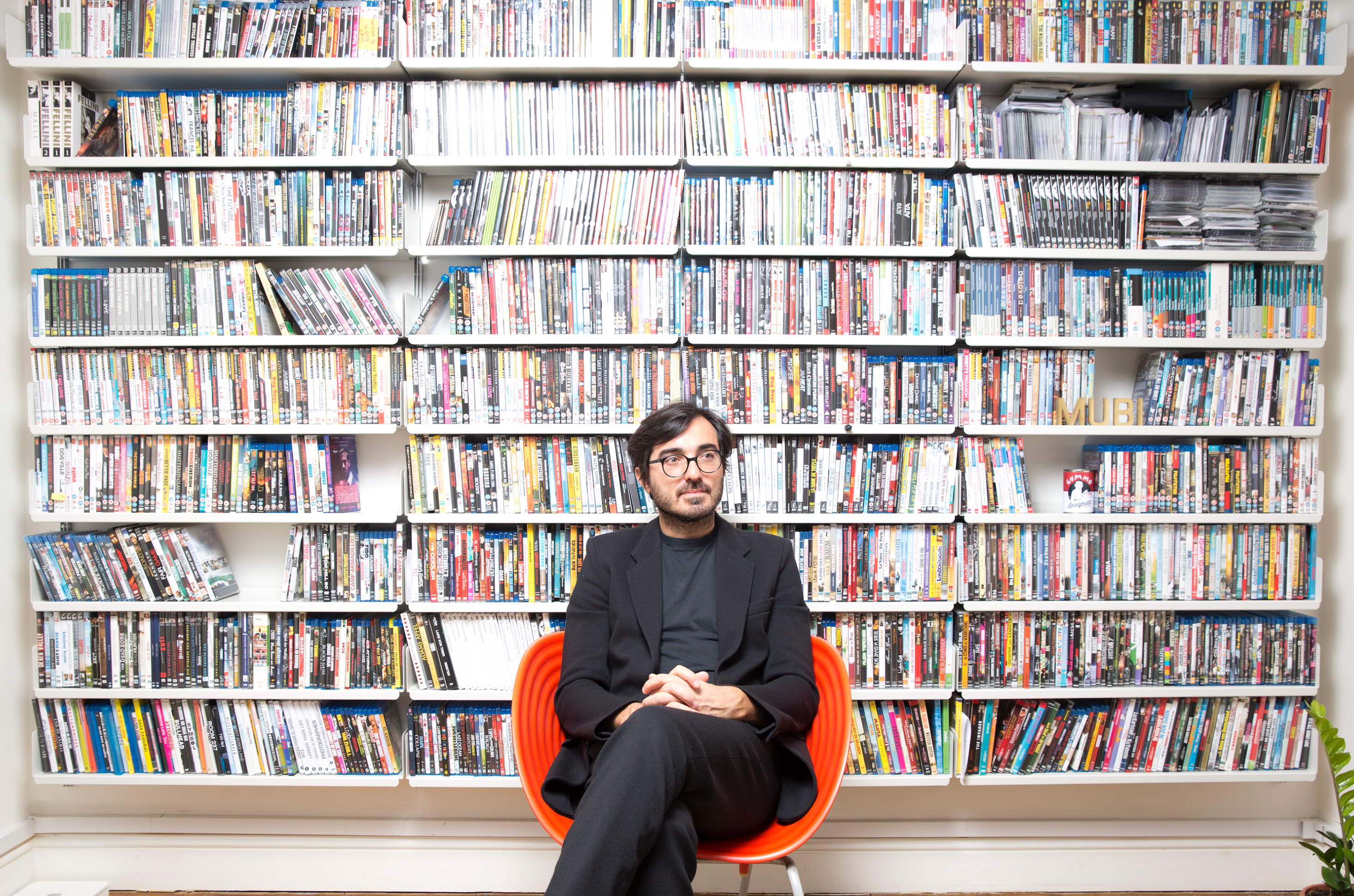Efe Çakarel seated in an orange chair in front of shelves filled with films
