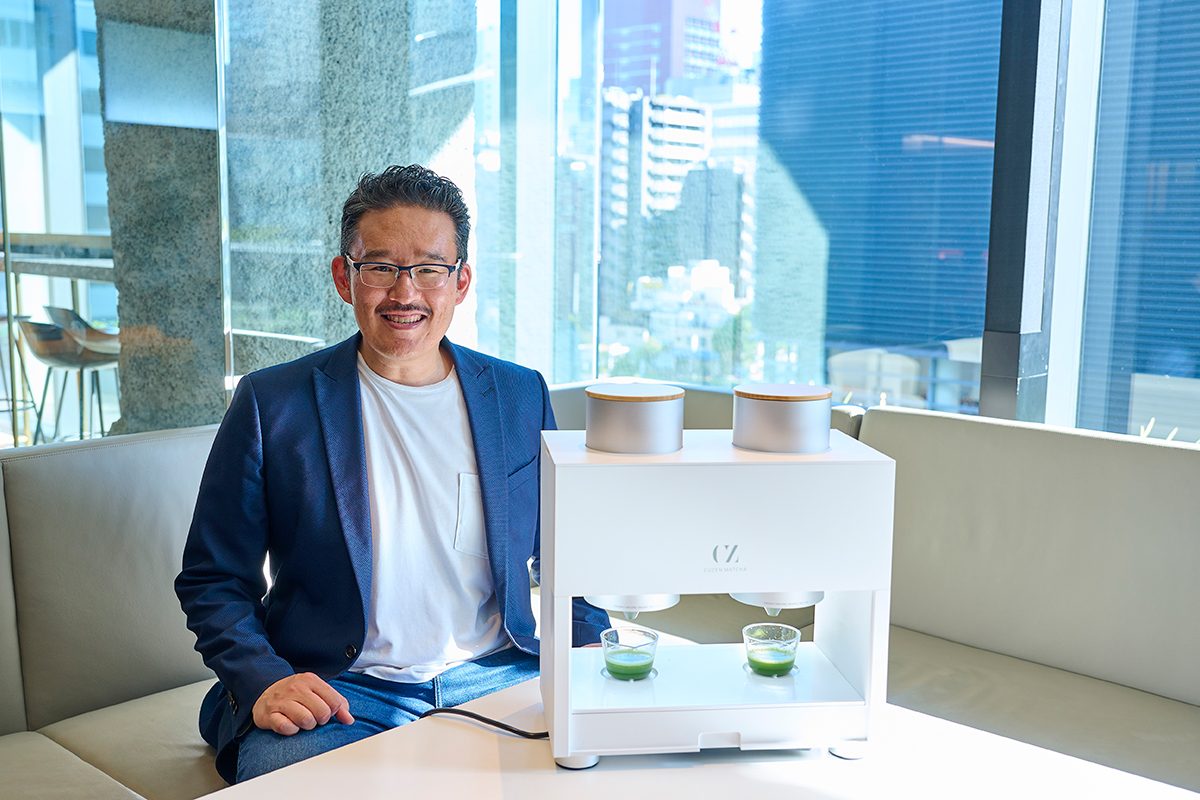 Eijiro Tsukada demonstrates the Cuzen Matcha Machine Pro beside two glasses of matcha