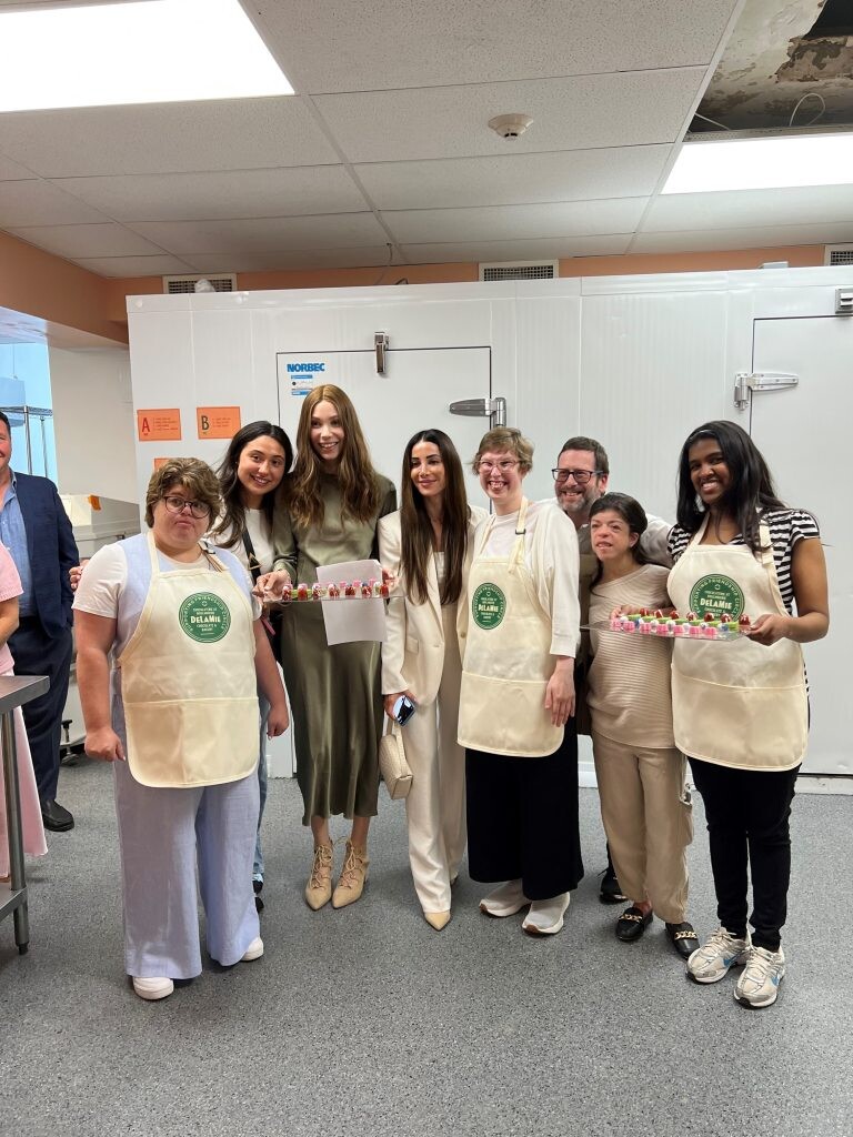 Elise Golbert in a white suit standing with the Friendship Circle Montreal team inside the DeLaMie chocolate workspace