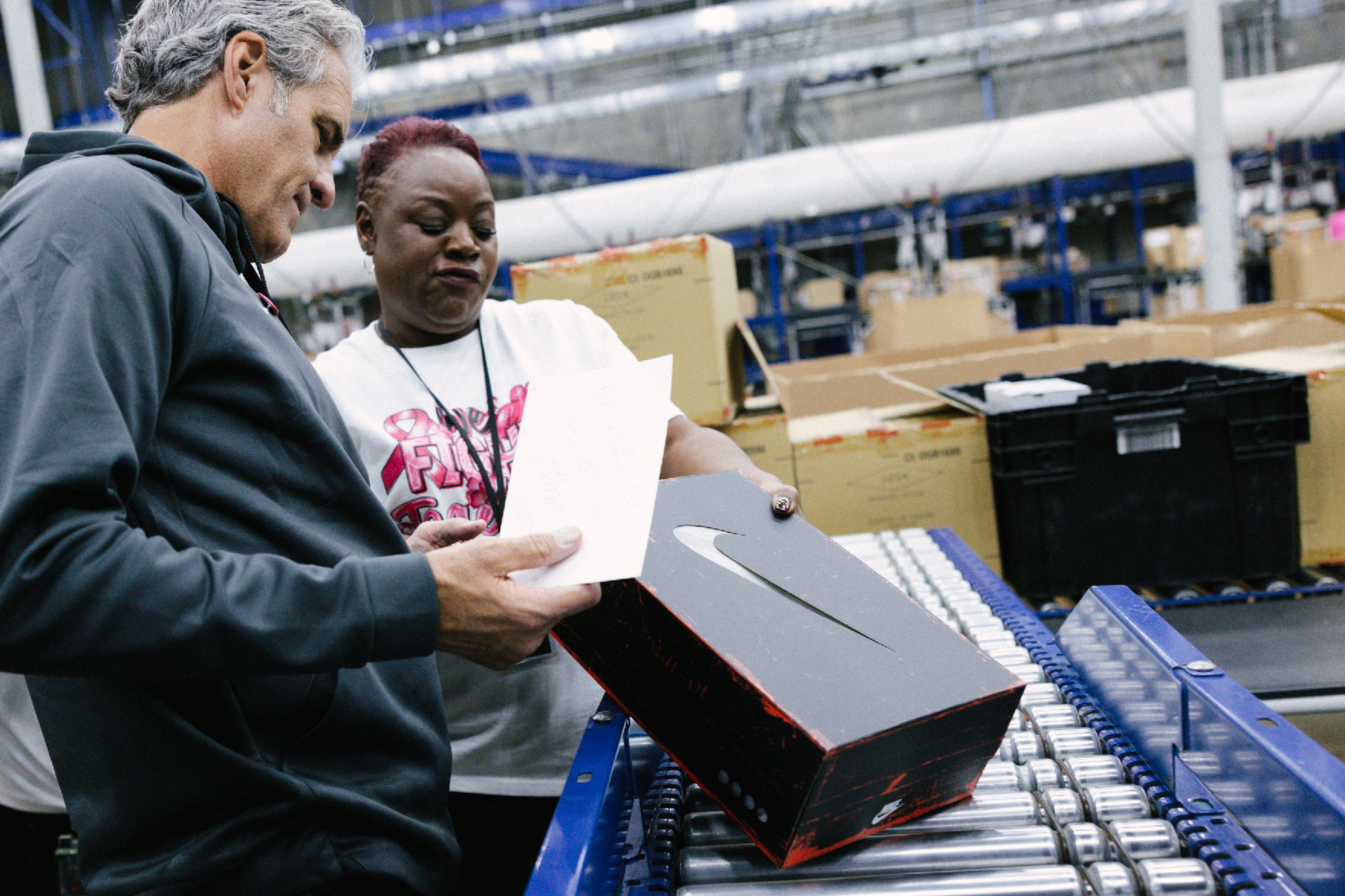 Elliott Hill and a Nike logistics employee beside a conveyor, holding a note and an Air Max shoe box