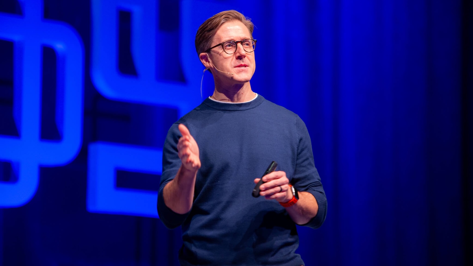 Elliott Parker wearing glasses and speaking onstage against a blue backdrop at Alloy’s 2024 venture-building summit