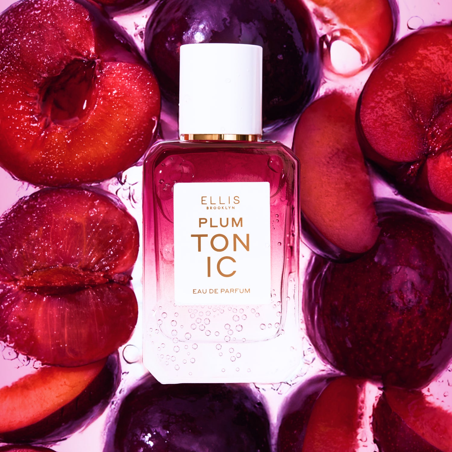 A burgundy PLUM TONIC perfume bottle surrounded by sliced plums underwater against a pink background