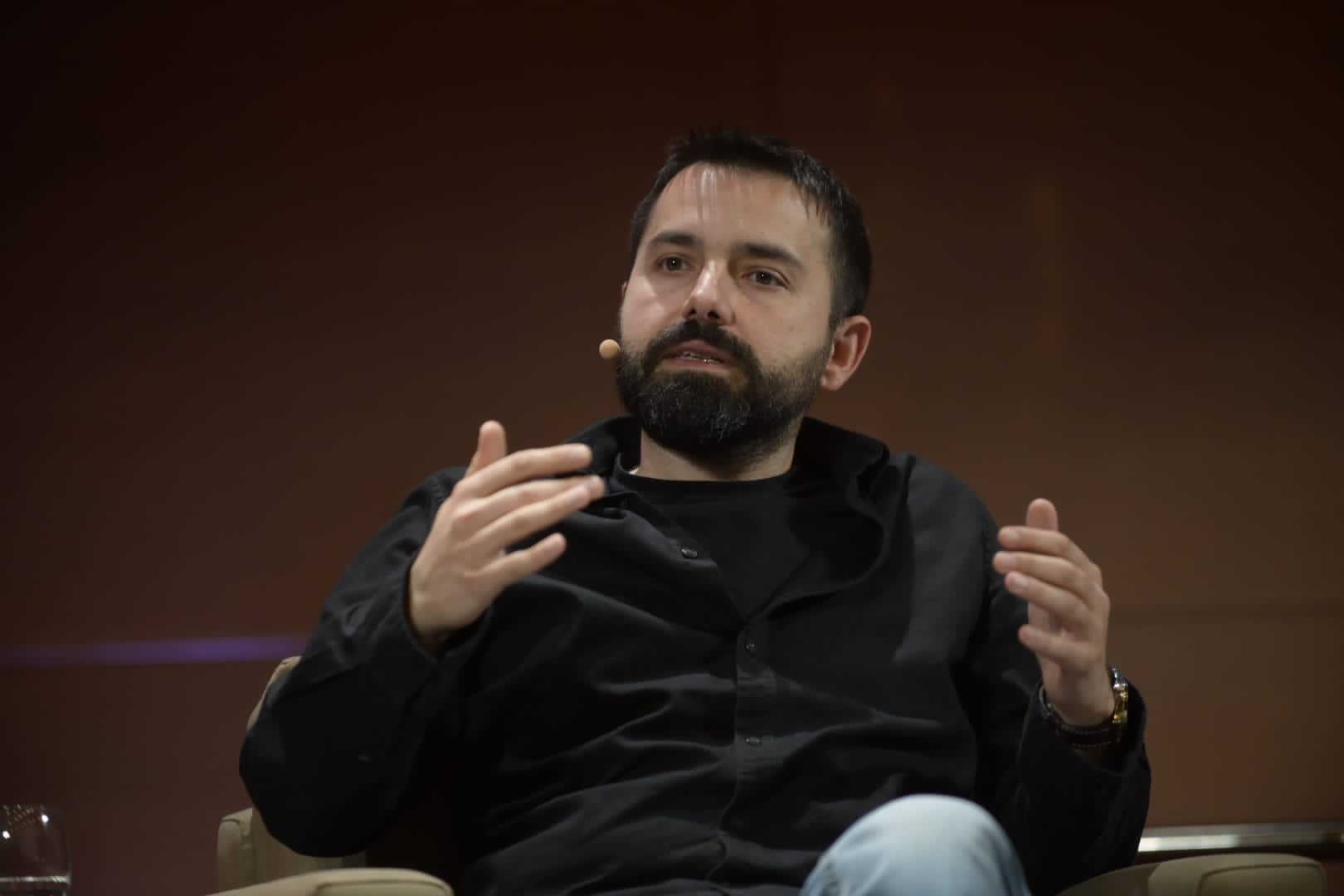 Emre Fadıllıoğlu seated with a headset microphone, gesturing during a speaking appearance