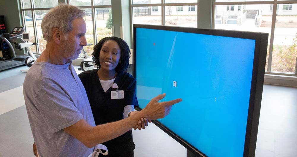 A patient practices an arm movement at a rehabilitation screen with a therapist beside him
