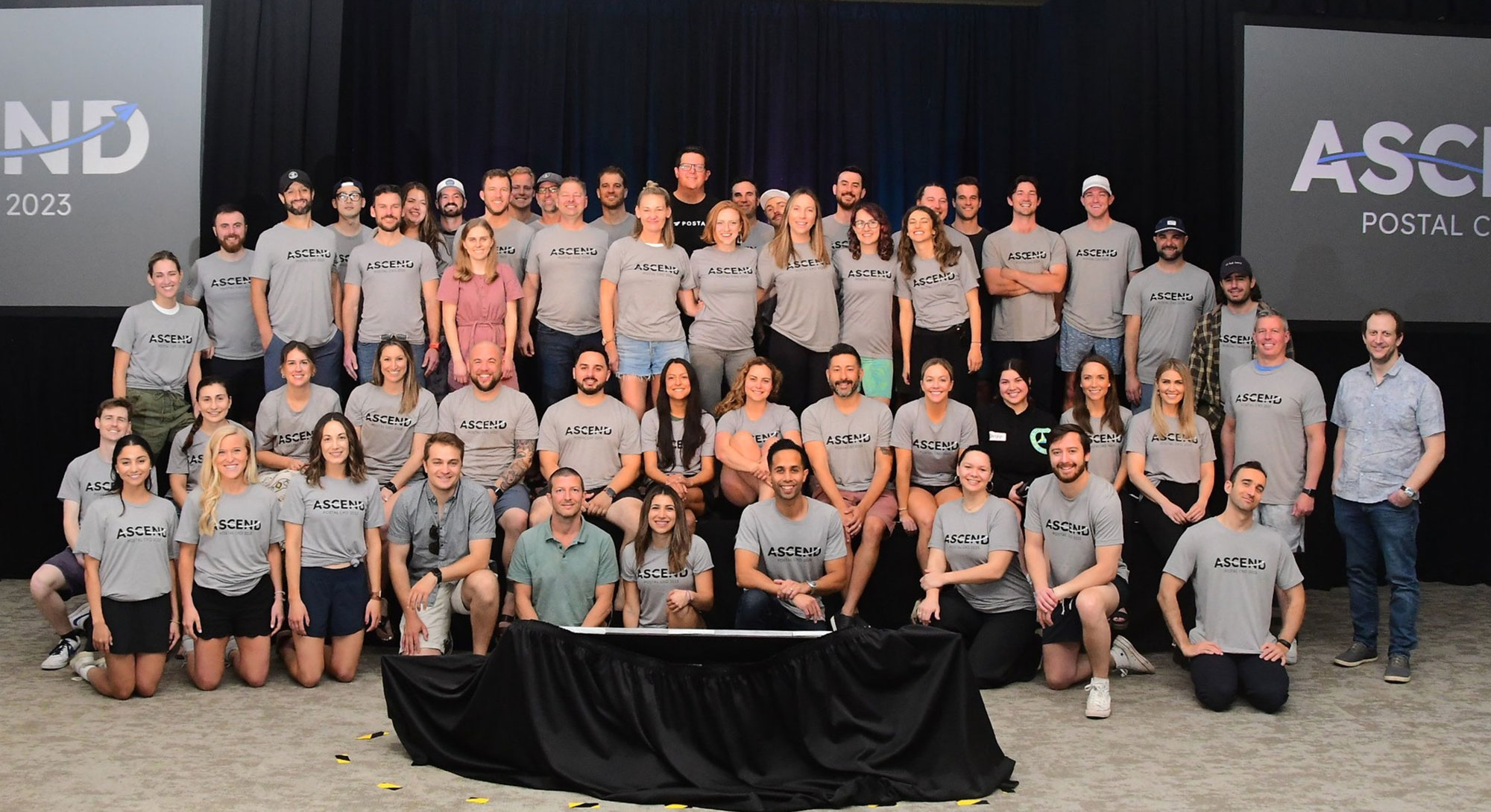 Erik Kostelnik in a black Postal shirt near the back center of a large Postal team group at Ascend 2023.