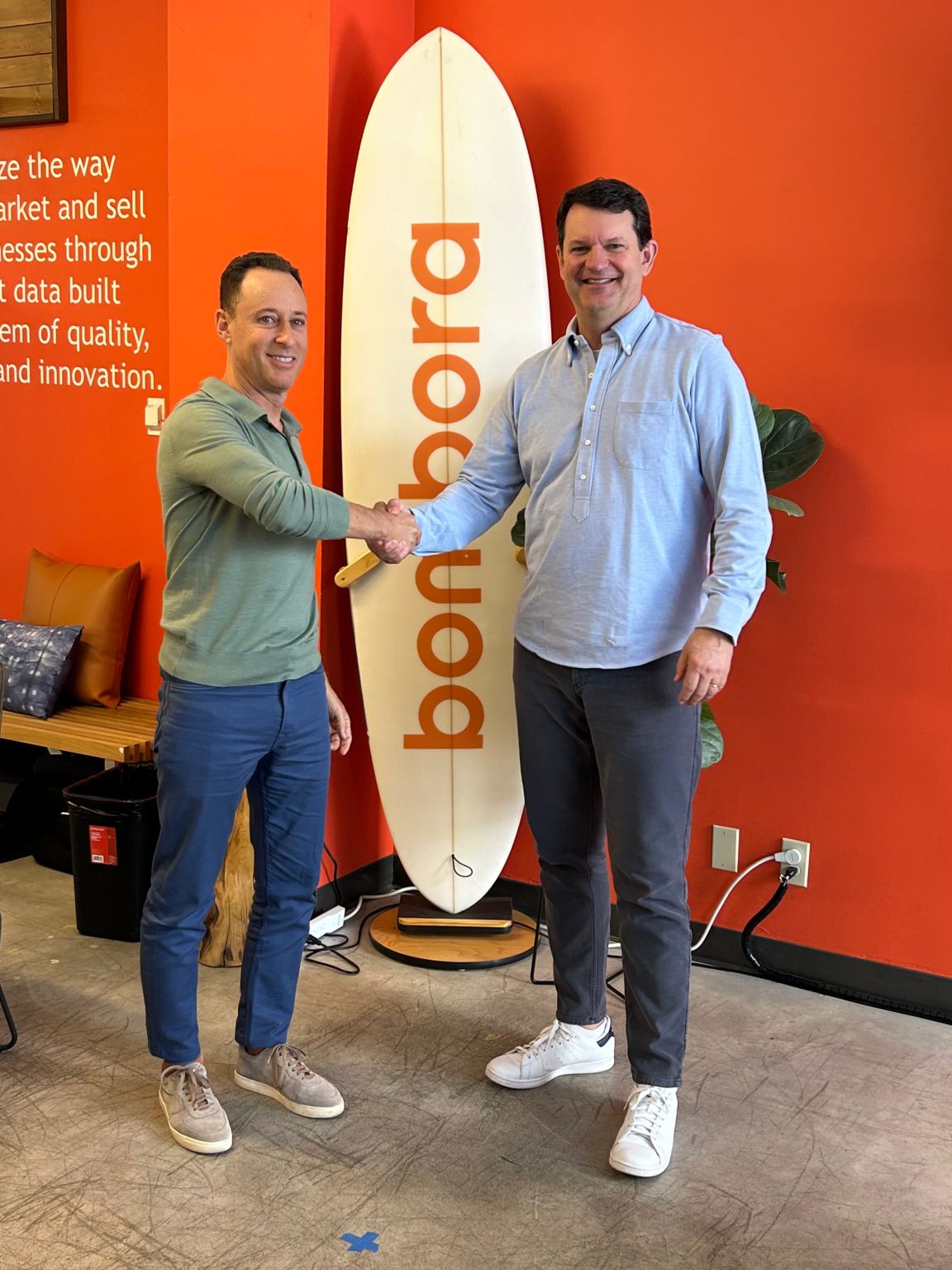 Erik Matlick, left, shaking hands with Mark Connon beside a Bombora surfboard