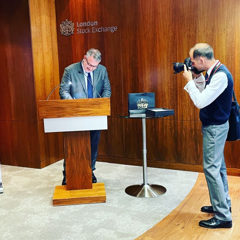 Erik Smith smiling at a lectern inside the London Stock Exchange while a photographer takes his picture