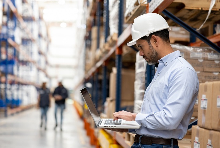 Warehouse worker in a hard hat using a laptop beside storage racks