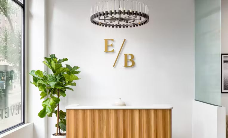 Ever/Body reception with a wood-fronted desk, plant and ceiling light