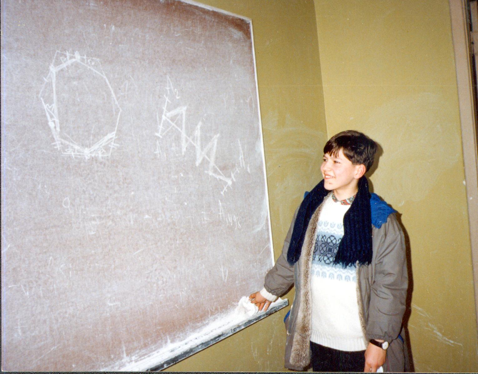 School-age Evgeny Sergeev explaining a mathematics solution beside a chalkboard