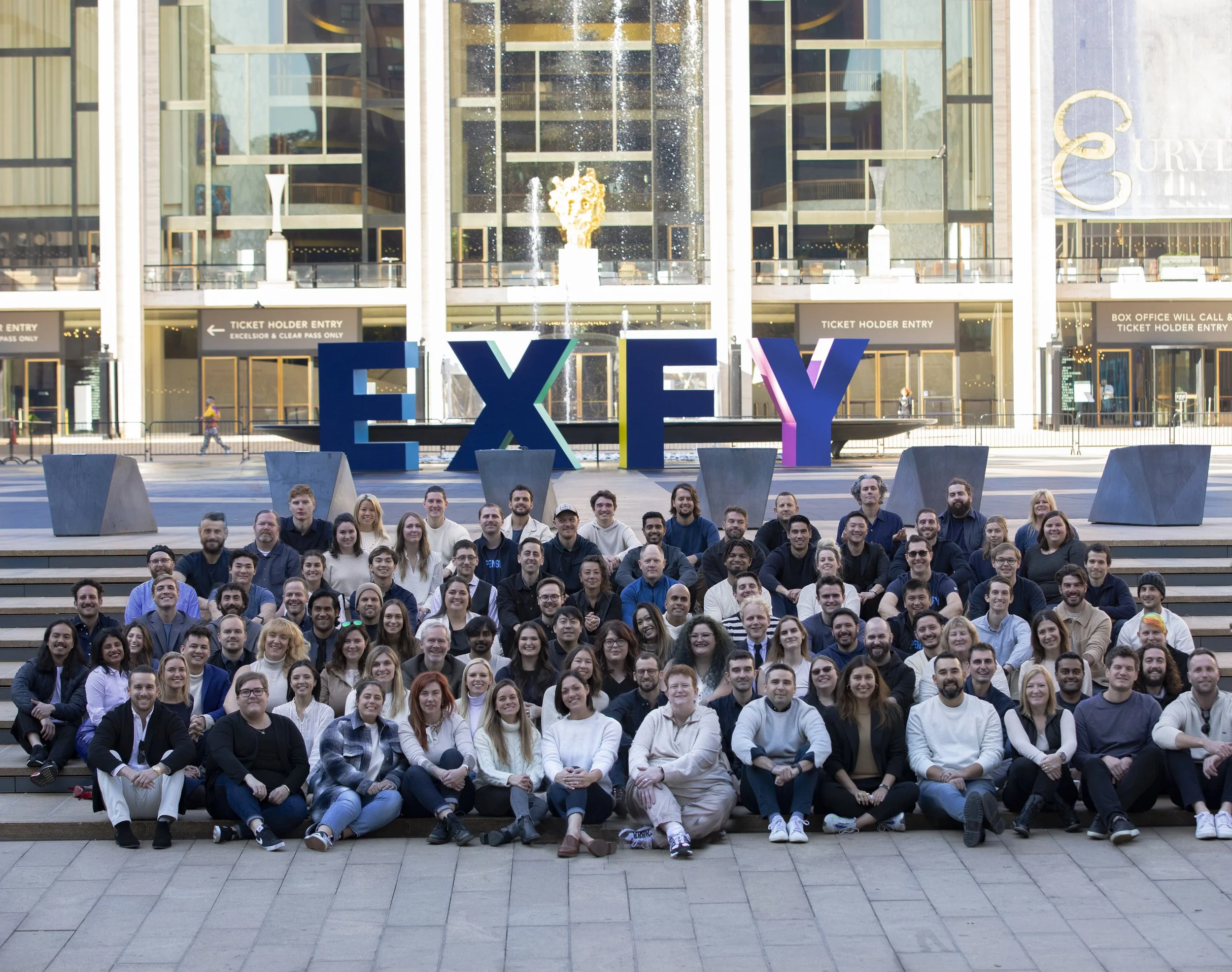 Expensify team gathered on outdoor steps beneath large EXFY letters