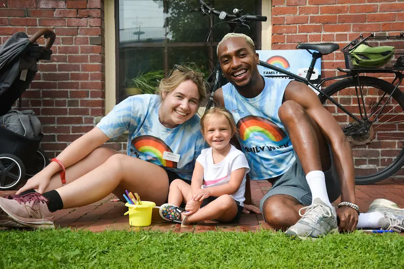 Two adults in Family Week shirts sitting outdoors with a child