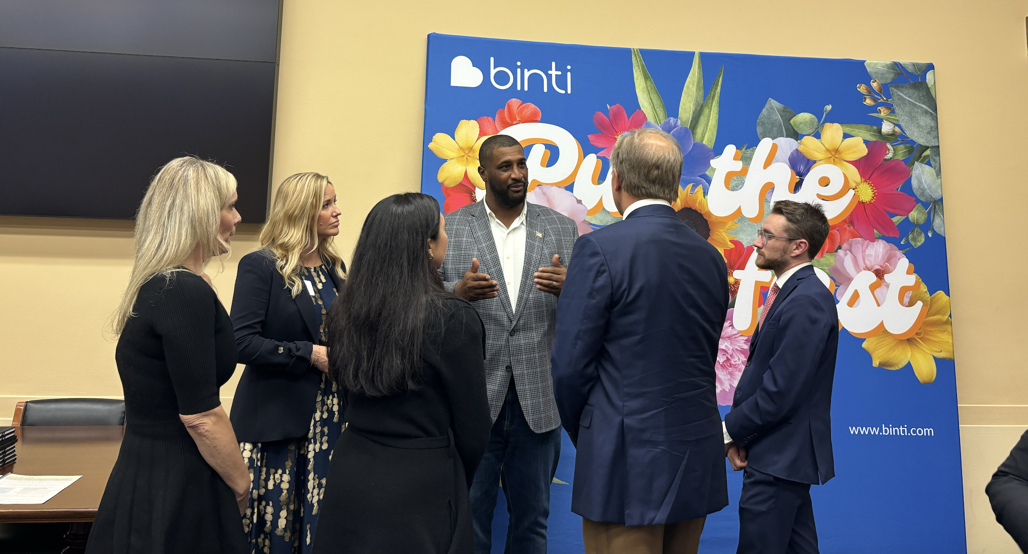 A small group, including Daunte Culpepper and Binti representatives, speaks in front of a colorful Binti display during a Capitol Hill gathering