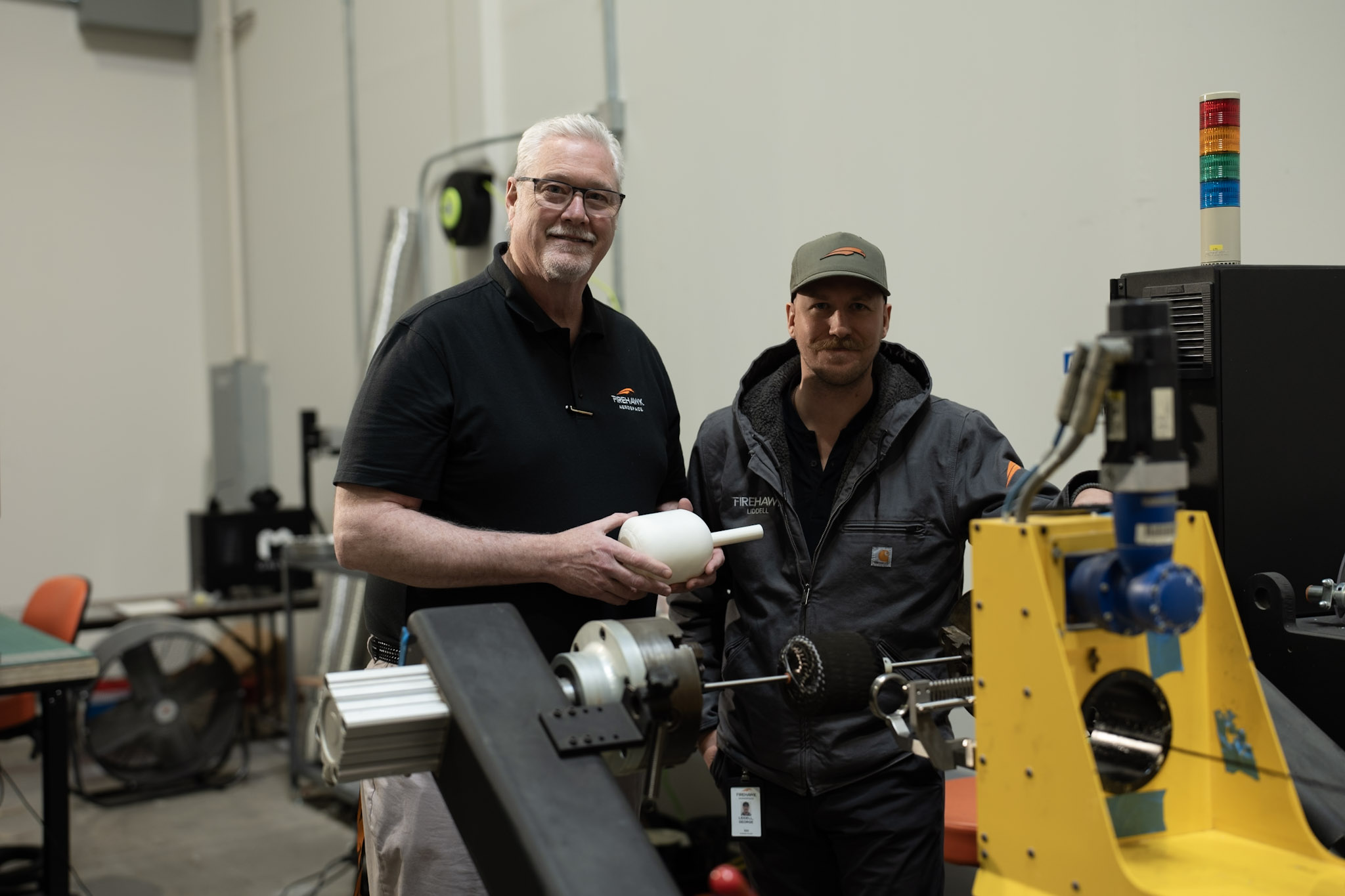 Two Firehawk team members stand beside manufacturing equipment while holding a white fuel-grain component