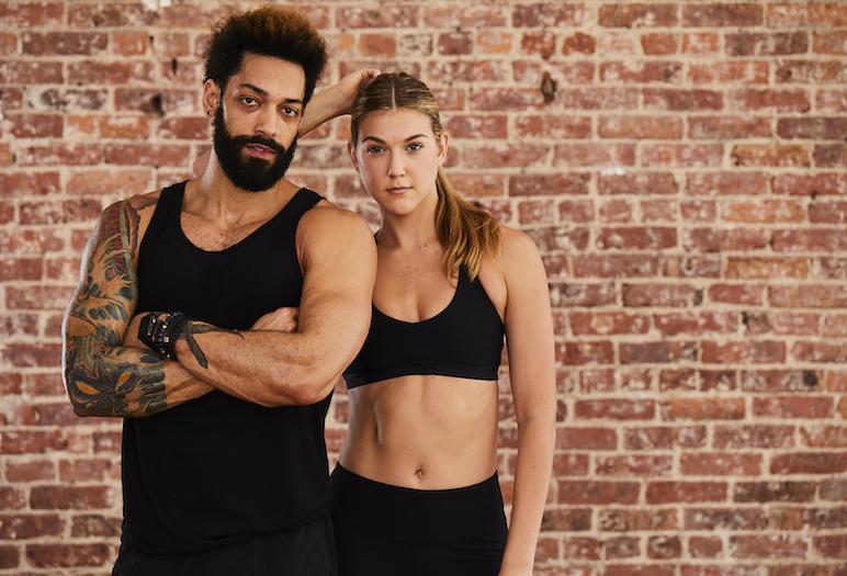 Two people in black workout clothes pose against a brick wall in a Fithouse press photograph