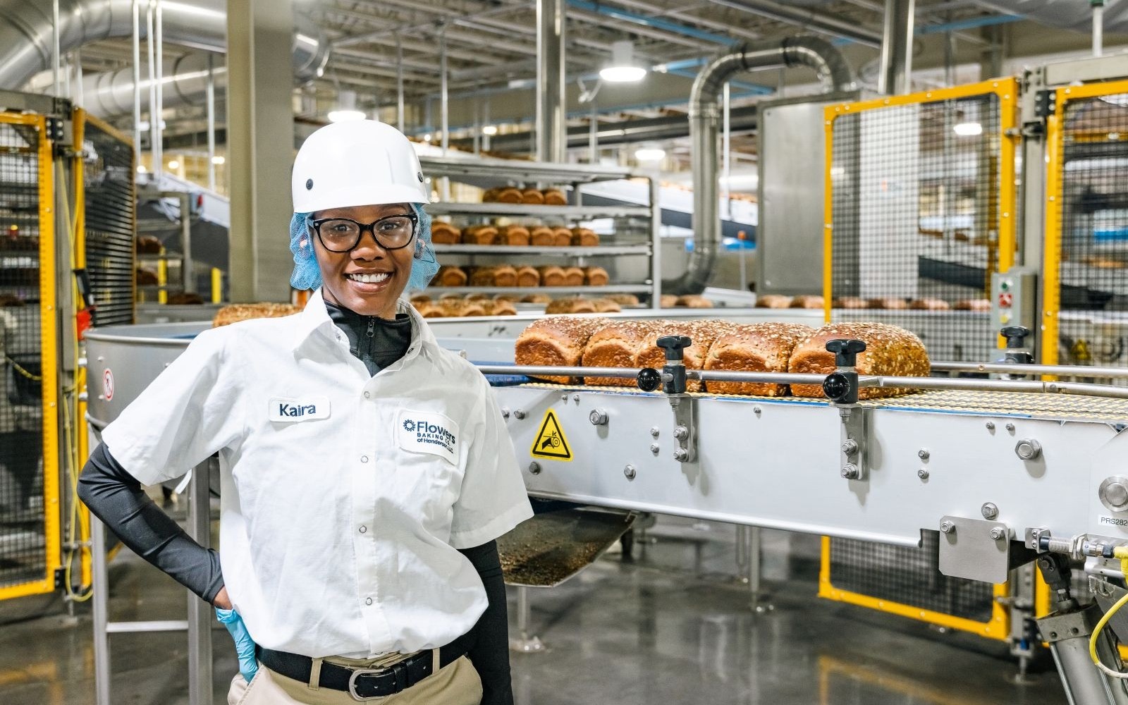 A Flowers bakery employee stands beside seeded loaves moving along a guarded production line.