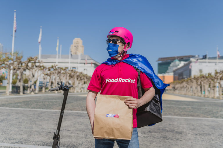 A Food Rocket courier in a pink uniform holding a grocery bag beside an electric scooter in San Francisco
