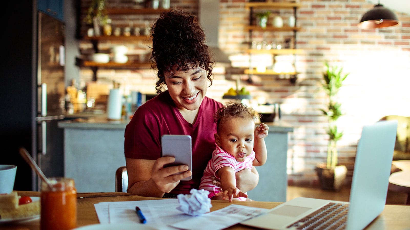 A woman holding a child looks at a phone beside a laptop and household paperwork
