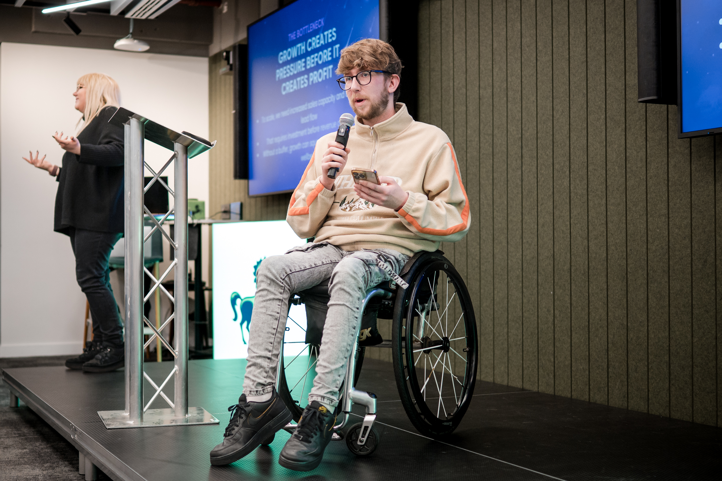 A seated speaker holds a microphone at Foundervine’s Breakthrough Demo Day, with another presenter alongside.