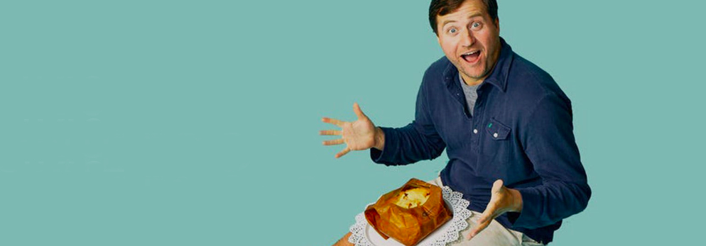 Frank Luciano smiling and presenting a wrapped food dish against a pale blue background