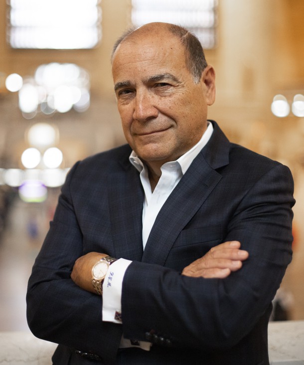 Frank Marino in a dark suit, standing with his arms folded in a public interior