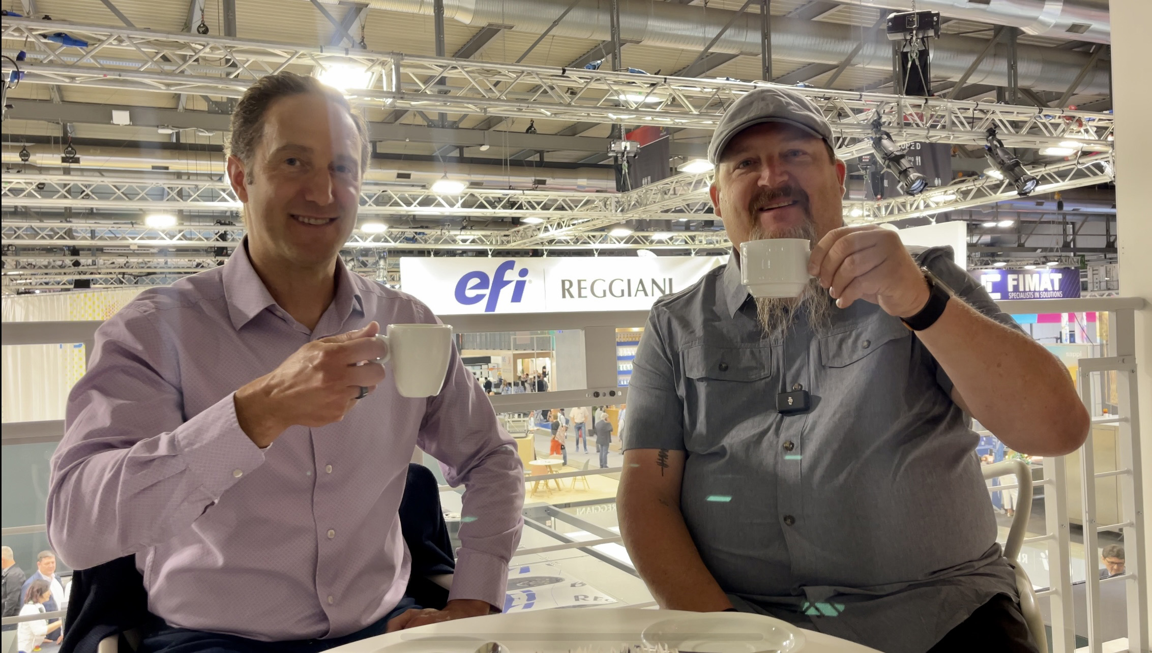 Frank Pennisi, left, and interviewer Eric Vessels holding coffee cups at an EFI exhibition stand
