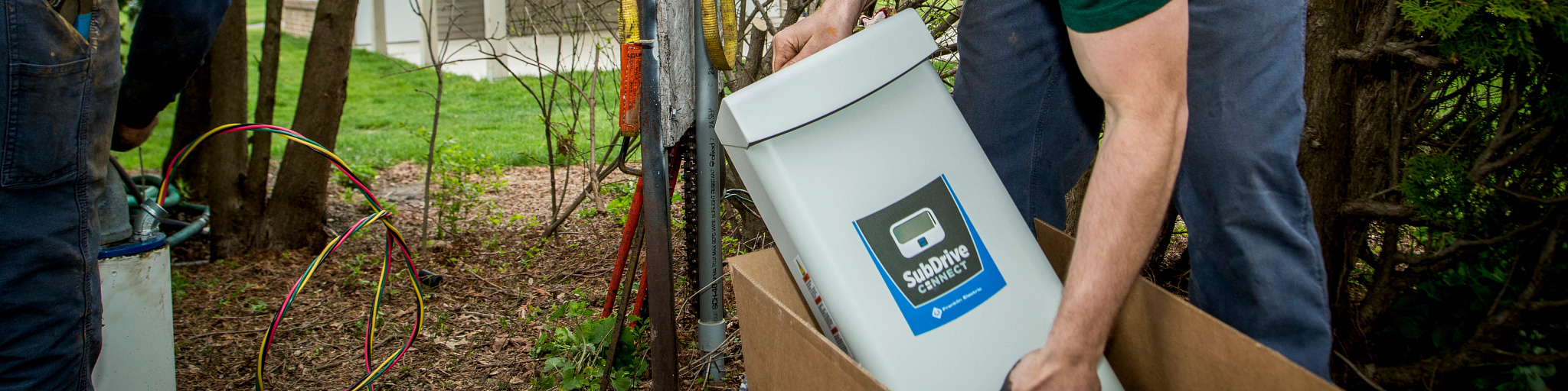 A technician lifts a SubDrive Connect control from its box beside a residential water-well installation