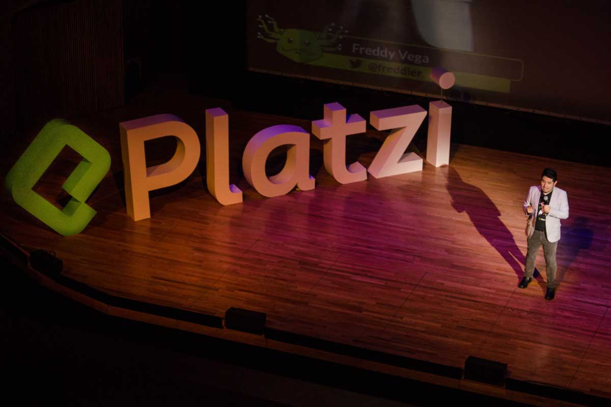 Freddy Vega speaking on a wooden stage beside large Platzi letters