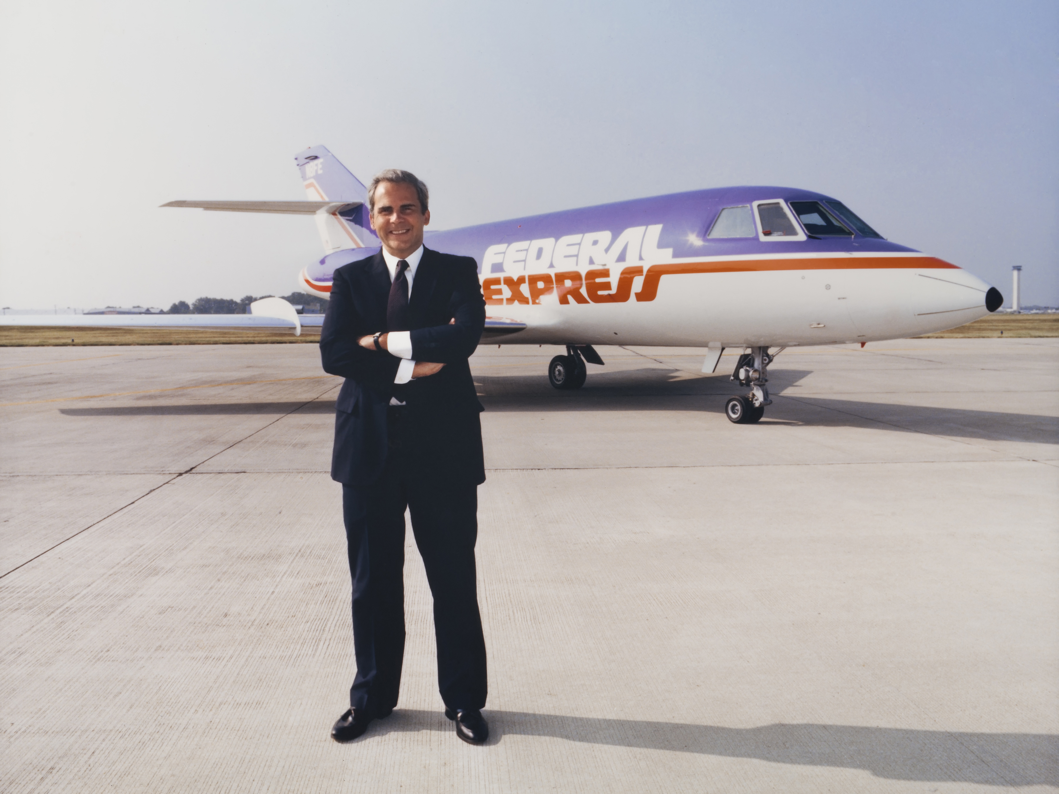 A young Fred Smith standing beside a purple, orange and white Federal Express Dassault Falcon jet