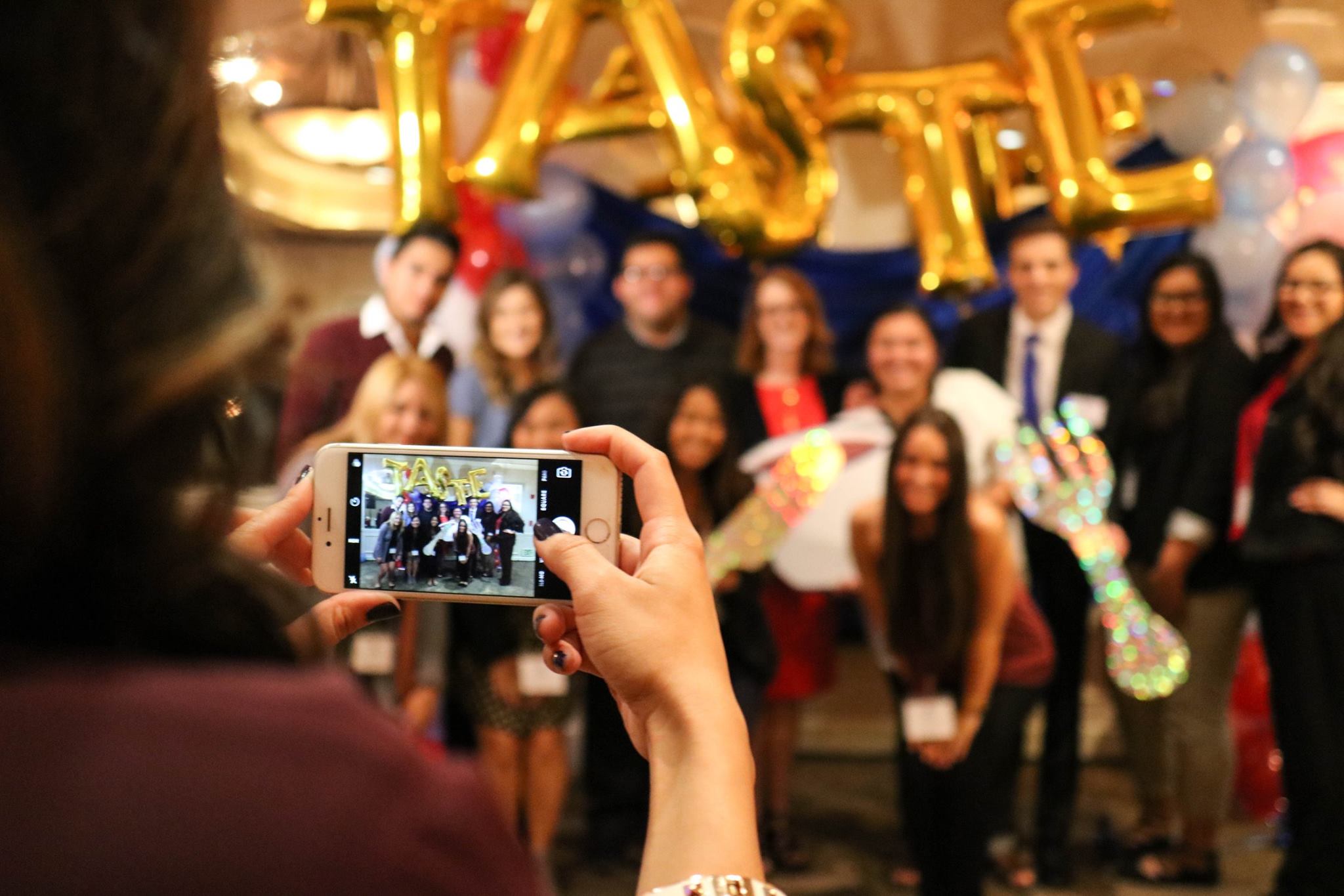 A phone frames a group of Fresno State students and faculty beneath gold TASTE balloons at the 2016 event.