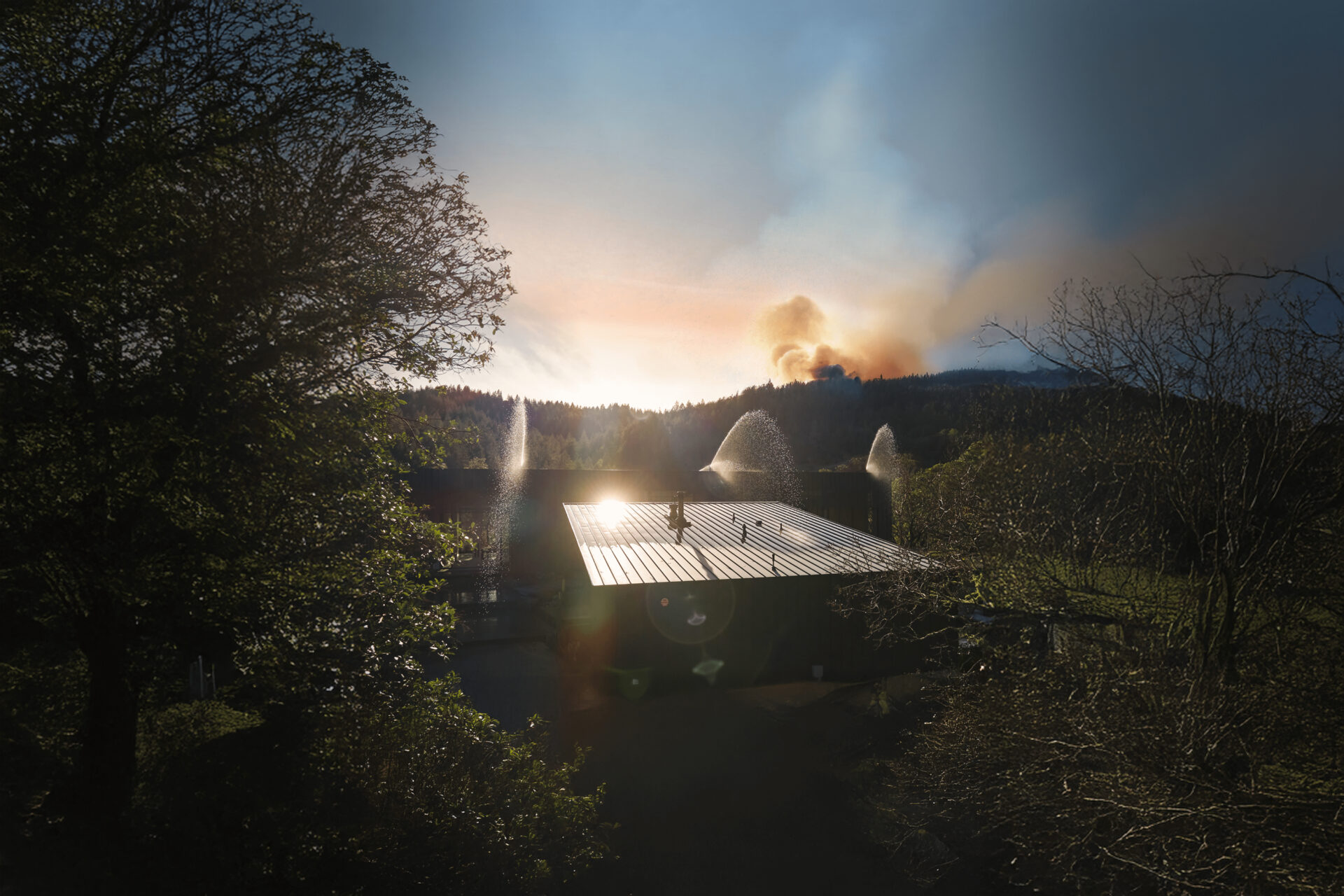 Exterior sprinklers operating on a home while smoke rises from a wildfire beyond a wooded ridge