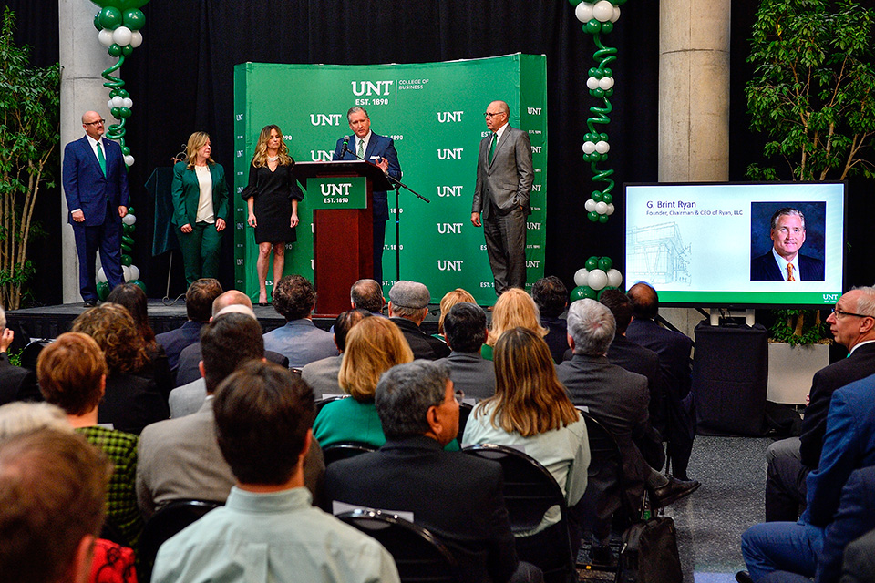 G. Brint Ryan speaking at a lectern during the UNT business college gift announcement, with guests watching.