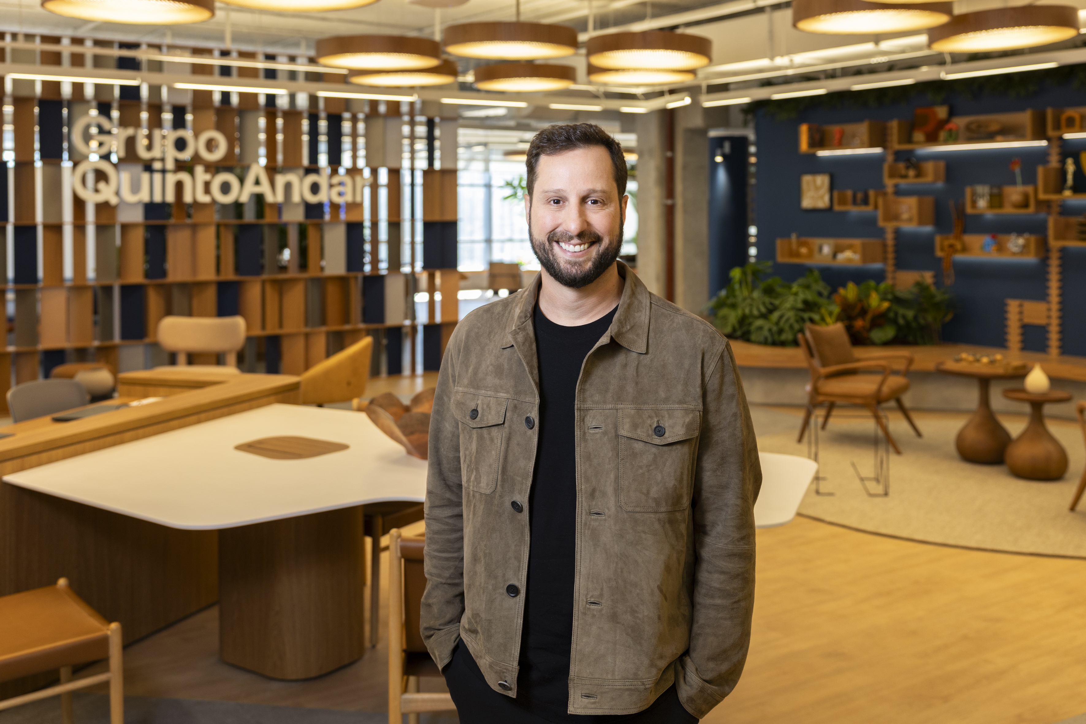 Gabriel Braga standing in a Grupo QuintoAndar office, with meeting tables and seating behind him
