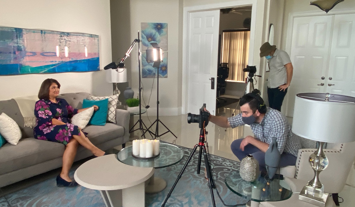 A masked video crew filming a seated interview subject in a living room
