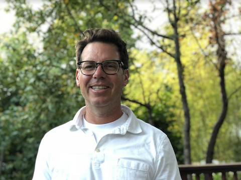 Gary Bird in glasses and a white shirt, standing outdoors among trees