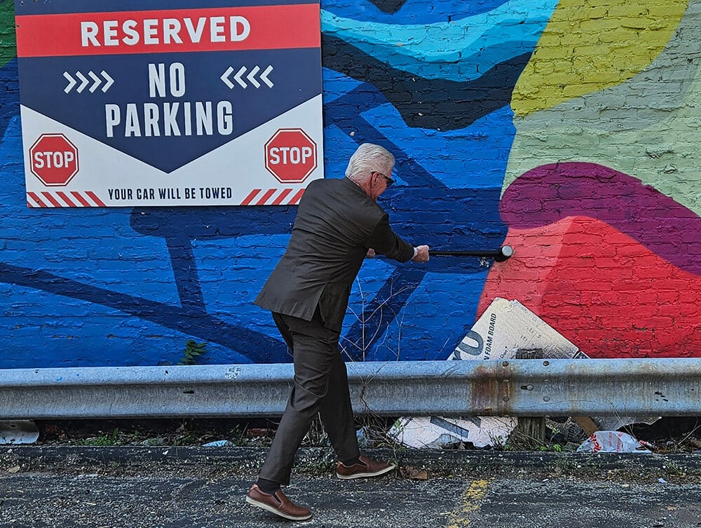 Gary Granger in a suit swinging a sledgehammer at the painted brick wall of the Walter Neller Building