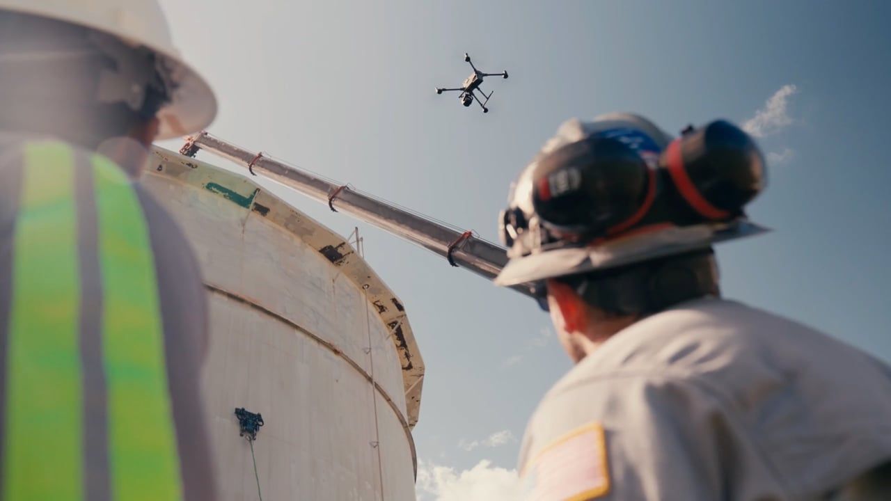 Two Gecko field engineers watch a drone near a storage tank while a crawler is attached to the tank wall