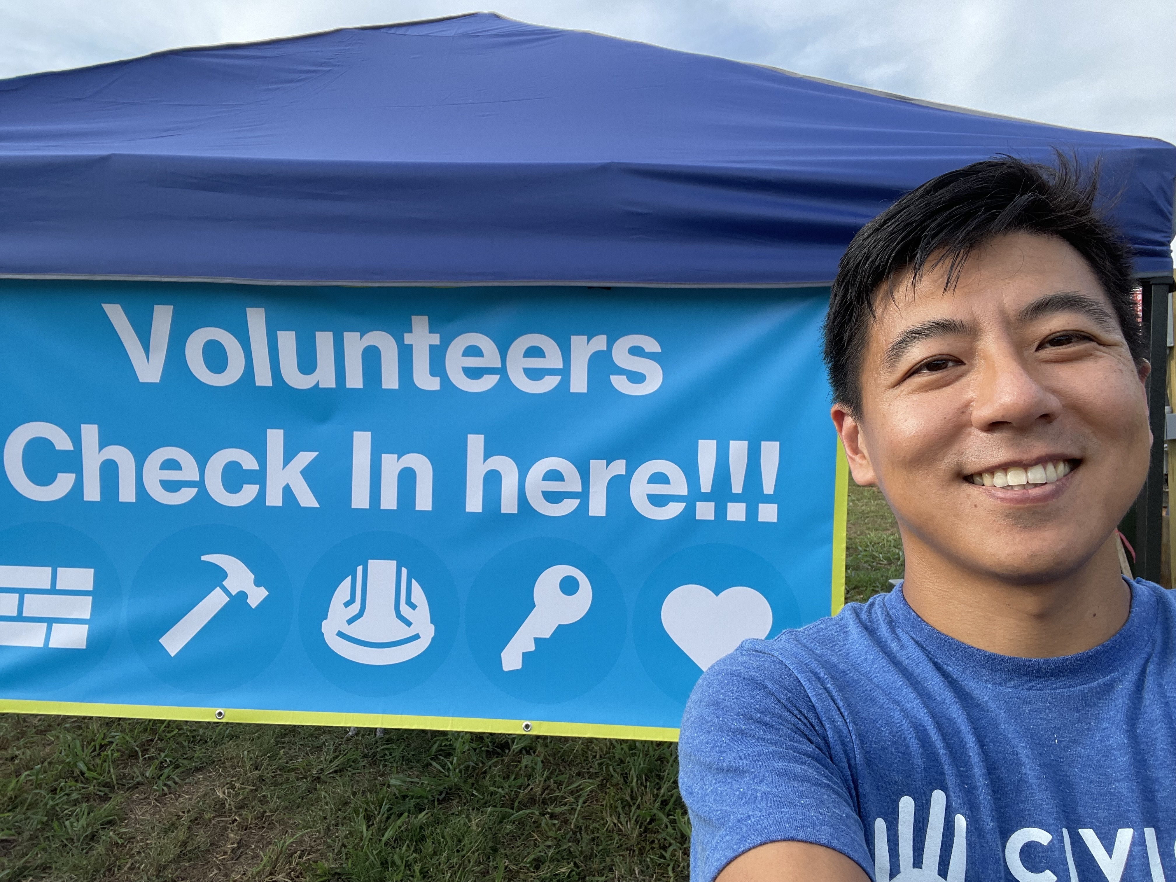 Geng Wang smiling beside a Volunteers Check In Here sign at a service event